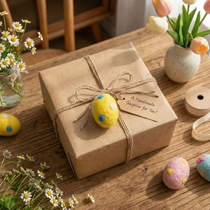 May include: A wrapped gift box tied with twine, adorned with a yellow felt egg with blue and white spots. A tag reads "A Handmade Surprise for You!". Additional decorative felt eggs and flowers are scattered on a wooden surface.