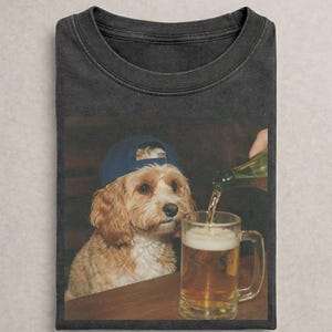 May include: A charcoal gray crew neck t-shirt featuring a graphic print of a light brown dog wearing a dark blue baseball cap. The dog is positioned next to a glass mug being filled with a golden amber liquid from a green glass bottle.