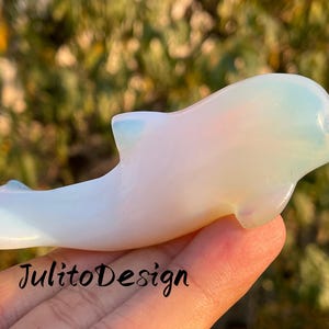 以下が含まれることがあります： 半透明でオパールのようなイルカの置物。彫刻は淡い白で、青とピンクの色合いがあり、手で持たれています。「JulitoDesign」の文字が画像の下部に表示されています。