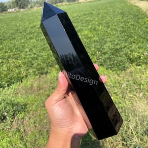 May include: A polished black obsidian obelisk, held in a hand, with a sharp, pointed top. The obelisk is rectangular with beveled edges and a glossy finish. The background is a field of green grass.