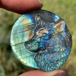 May include: A round, iridescent labradorite stone carved with a wolf's head. The stone features shades of blue, green, and gold. The word "Design" is visible on the stone. The carving is detailed and the stone is held by a person.