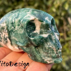 May include: A carved skull made of green and white stone. The skull has detailed features, including eye sockets, a nose cavity, and a jawline. The surface of the skull is polished and has a marbled appearance. The text "JulitoDesign" is visible.