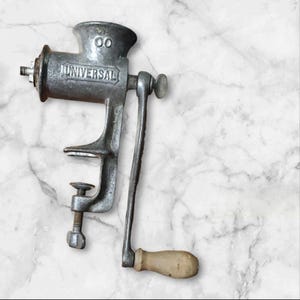 May include: A vintage, silver-colored Universal meat grinder with a hand crank and wooden handle. The grinder is clamped to a surface and features the word "UNIVERSAL" embossed on its side. The top of the grinder has the number "00".