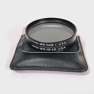 May include: A Tiffen 58mm camera lens filter with the text "Tiffen 58M Haze 1 U.S.A." and "Tiffen 58M ND 0.6 U.S.A." The filter is black and sits on a black leather case.