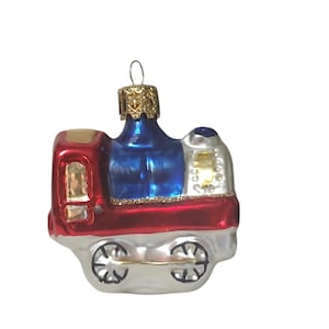 May include: A glass train ornament featuring a shiny red front section, a deep blue central boiler, and a silver base with black-outlined wheels. The top includes a gold-colored metal cap with a small loop for hanging, and gold accents.