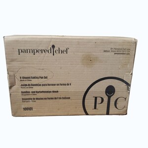 May include: A cardboard box for a Pampered Chef V-Shaped Baking Pan Set. The box has the Pampered Chef logo and product information printed on it. The box is beige with black text and a black circular logo.
