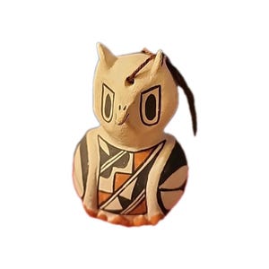 Puede incluir: Figura de búho de cerámica de color crema, con grandes ojos negros y un pico pequeño. El pecho del búho presenta un diseño geométrico en negro, naranja y blanco. Una cuerda marrón está unida en la parte superior para colgar.