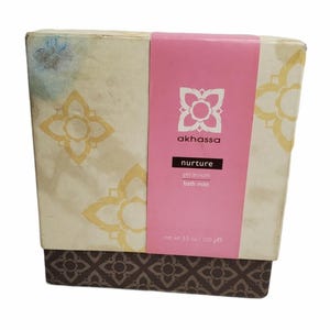 May include: A rectangular box with a pink band and floral designs. The box has a cream-colored background with gold floral patterns. The pink band features the brand name "akhassa" and the word "nurture". The box contains a geranium bath mitt and weighs 3.5 oz / 100 g.