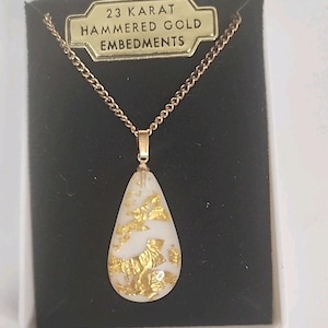 Könnte beinhalten: Goldfarbene Halskette mit tropfenförmigem Anhänger. Der Anhänger ist weiß mit Goldblatt-Akzenten. Die Halskette wird in einer schwarzen Schachtel mit einem goldenen Etikett mit der Aufschrift "23 Karat Hammered Gold Embedments" präsentiert.
