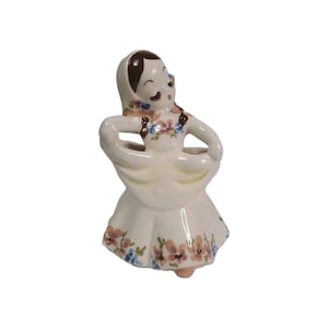 May include: A vintage ceramic figurine of a woman in a dress, with her hands on her hips. The figurine is painted in cream, with brown hair and floral accents in pink and blue. The woman's eyes are closed, and she has a serene expression.