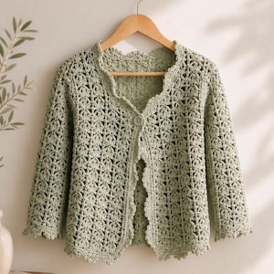 K&ouml;nnte beinhalten: Hellgr&uuml;ne H&auml;keljacke mit gewelltem Saum und einem einzelnen Knopfverschluss. Die Dreiviertel&auml;rmel und das zarte Blumenmuster erzeugen eine feminine und luftige &Auml;sthetik. Die Strickjacke wird auf einem Holzb&uuml;gel pr&auml;sentiert.