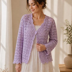 K&ouml;nnte beinhalten: Ein helllila H&auml;kelcardigan mit gewelltem Saum und einem einzelnen Knopfverschluss. Der lang&auml;rmlige, offen gewebte Pullover wird &uuml;ber einem wei&szlig;en Oberteil und einem langen, flie&szlig;enden Rock getragen. Das zarte Muster und die weiche Farbe erzeugen eine feminine &Auml;sthetik.