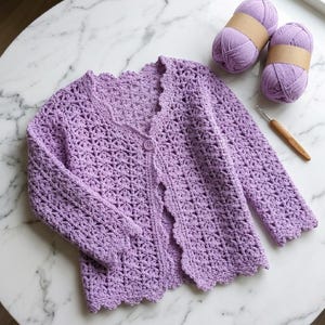 K&ouml;nnte beinhalten: Ein helllila geh&auml;kelter Cardigan mit gewelltem Rand und einem einzelnen Knopfverschluss. Zwei Kn&auml;uel passende Wolle und eine H&auml;kelnadel liegen neben dem Cardigan auf einer Marmoroberfl&auml;che. Der Cardigan hat ein Blumenmuster.