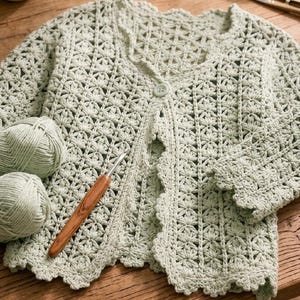 K&ouml;nnte beinhalten: Hellgr&uuml;ne H&auml;keljacke mit floralem Muster und einem einzelnen Knopfverschluss. Die Jacke wird mit zwei Kn&auml;ueln passendem Garn und einer H&auml;kelnadel gezeigt. Die Kanten der Jacken haben eine gewellte Bord&uuml;re.