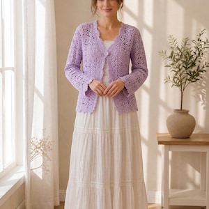 K&ouml;nnte beinhalten: Ein helllila H&auml;kelcardigan mit gewelltem Saum und Knopfverschluss. Der Cardigan wird mit einem wei&szlig;en, mehrlagigen Maxirock kombiniert. Das Outfit ist f&uuml;r Fr&uuml;hling oder Sommer geeignet.