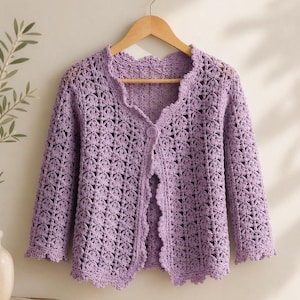 K&ouml;nnte beinhalten: Ein helllila H&auml;kelcardigan mit floralem Muster. Der offene Cardigan hat einen gewellten Saum und wird mit einem einzelnen Knopf geschlossen. Die &Auml;rmel sind dreiviertellang. Der Cardigan h&auml;ngt an einem Holzb&uuml;gel.
