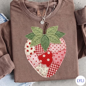 May include: A brown sweatshirt with a large, appliqued strawberry design. The strawberry is made of various fabric patterns, including red gingham, floral, and heart prints. The strawberry has green leaves and a brown stem. A silver necklace is draped over the sweatshirt.
