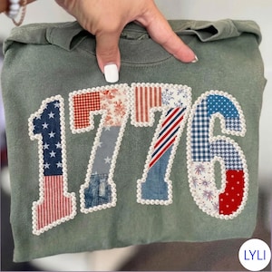 May include: Olive green sweatshirt with the number "1776" appliqued on the front. The numbers are made of various patriotic patterns, including stars, stripes, and floral designs, and are outlined with a pearl trim. The sweatshirt is held by a hand.