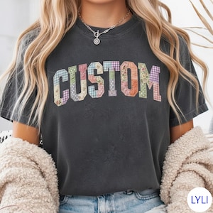 May include: Dark gray t-shirt with the word "CUSTOM" in a patchwork design. The letters are made of various floral and plaid patterns in pastel colors. The shirt has a vintage, worn look.