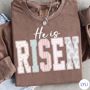 May include: A brown Comfort Colors sweatshirt with the words "He is Risen" in white script and patchwork letters. The letters are decorated with floral, polka dot, and striped patterns in pastel colors. A silver necklace hangs on the neckline.
