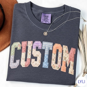 May include: Dark gray t-shirt with the word "CUSTOM" in patchwork letters. The letters are made of various floral and patterned fabrics. A silver necklace with a pendant is on the shirt. The shirt has a Comfort Colors tag.