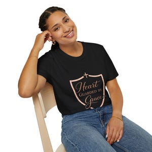 May include: Black t-shirt featuring a shield design with the words "Heart Guarded by Grace" in elegant script. The shield and text are in a rose gold color. The shirt is made of a soft material.