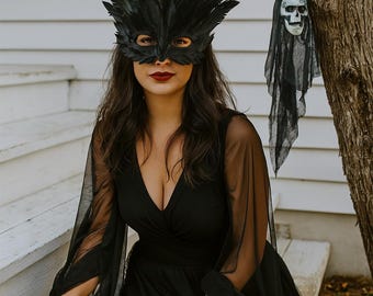 May include: A black feather mask with eye cutouts. The mask is worn with a black dress with sheer sleeves. A decorative skull hangs from a tree branch in the background. The woman has dark hair and red lipstick.
