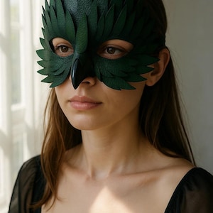 May include: A dark green leather mask in the shape of a bird's face, with detailed feather-like cutouts. The mask covers the eyes and nose, with a beak-like extension. The person is wearing a black top.