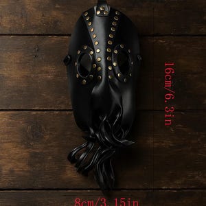 May include: A black plague doctor mask with brass rivets and eye holes. The mask is 6.3 inches (16 cm) tall and 3.15 inches (8 cm) wide. The mask has a beak-like nose and a strap for wearing.