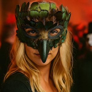 May include: A green and black feather mask with a bird-like beak. The mask covers the eyes and nose, with a crown of green feathers. The person has blonde hair and is wearing a black top. The mask is likely for a costume or masquerade.