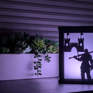 May include: A decorative black framed light box featuring a silhouette of a medieval knight and castle. The light box is on a wooden surface next to a white rectangular planter filled with succulents. The scene is illuminated with a soft purple glow.