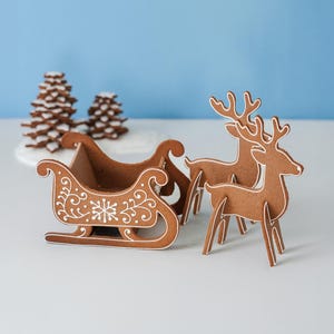 May include: A brown gingerbread-style sleigh and two standing reindeer figures with white icing-like decorative scrollwork and snowflake patterns. In the background, two stacked gingerbread-style pine trees are visible on a white base.