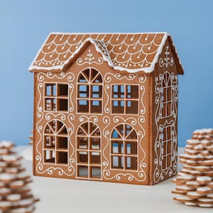 May include: A brown gingerbread house with white icing details, featuring arched windows and a shingled roof design. Two stacked cookie trees with white icing accents are positioned in the foreground, flanking the structure on both sides.