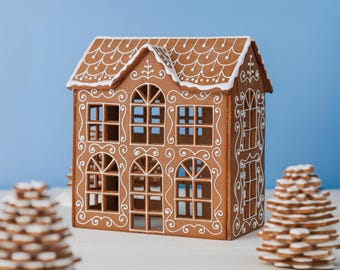 May include: A brown gingerbread house with white icing details, featuring arched windows and a shingled roof design. Two stacked cookie trees with white icing accents are positioned in the foreground, flanking the structure on both sides.