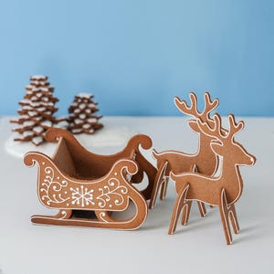 May include: Gingerbread Christmas decorations featuring a sleigh and reindeer. The brown gingerbread pieces are decorated with white icing details. Two gingerbread Christmas trees are in the background against a blue backdrop.