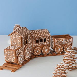 Może przedstawiać: Piernikowy pociąg z misternymi białymi zdobieniami z lukru, w tym dekoracyjnymi kołami i wzorami na dachu, stoi na drewnianym torze. Dwa piernikowe choinki znajdują się na pierwszym planie. Tło jest jasnoniebieskie.