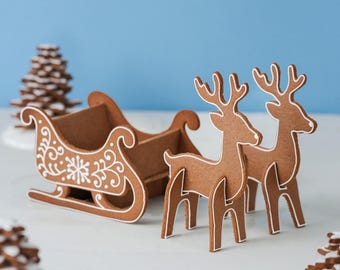 May include: A brown gingerbread-style sleigh and two standing reindeer figures, each featuring white piped icing details. The sleigh has a decorative snowflake and scroll pattern on its side, while the reindeer have white outlines on their bodies.