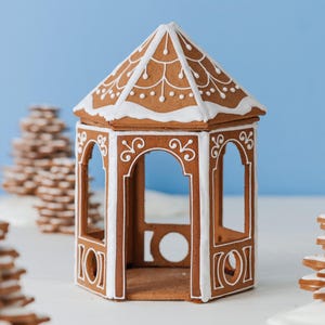 May include: A brown gingerbread gazebo with a hexagonal roof and open arched sides. The structure is decorated with white icing in intricate scrollwork patterns along the roof panels and pillars, featuring a pointed roof and a hollow interior.