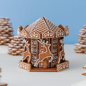 May include: A gingerbread carousel structure featuring brown cookie panels decorated with white icing. The roof has a cross-hatched pattern, and the sides display white icing silhouettes of reindeer and intricate scrollwork designs.