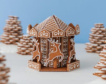 May include: A gingerbread carousel structure featuring brown cookie panels decorated with white icing. The roof has a cross-hatched pattern, and the sides display white icing silhouettes of reindeer and intricate scrollwork designs.