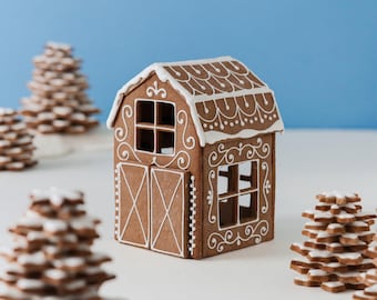 Op de afbeelding: Een peperkoekschuur met bruine koekjesmuren versierd met witte glazuurkrullen, een zadeldak met witte glazuurshingles en open raamkozijnen, omringd door kleine gestapelde peperkoekkoekjes in de vorm van bomen.