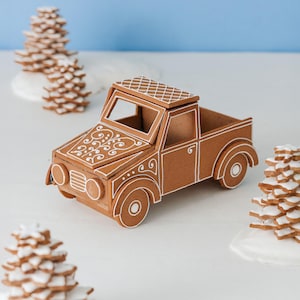 Könnte beinhalten: Ein brauner Lebkuchen-LKW mit weißem Zuckerguss auf der Motorhaube, dem Dach und der Karosserie, umgeben von drei kleinen gestapelten sternförmigen Keksbäumen mit weißem Zuckerguss auf einer glatten Fläche. Der LKW hat runde Scheinwerfer.