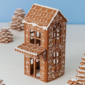 Könnte beinhalten: Ein braunes Lebkuchenhaus-Modell mit weißer Glasurverzierung an Dach und Wänden. Das Haus hat ein offenes Rahmendesign mit Bogenfenstern, einem kleinen Vordach und filigranen weißen Schnörkelmustern, die die Außenfassade schmücken.