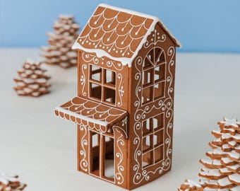 May include: A brown gingerbread house structure featuring white icing details on the roof and walls. The house has an open-frame design with arched windows, a small front awning, and intricate white scrollwork patterns decorating the exterior.