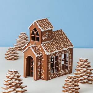 Può includere: Una casa di pan di zenzero con dettagli in glassa bianca e diversi alberi di pan di zenzero. La casa ha un tetto dettagliato, finestre e un ingresso ad arco. Gli alberi sono fatti di biscotti di pan di zenzero impilati con glassa bianca.