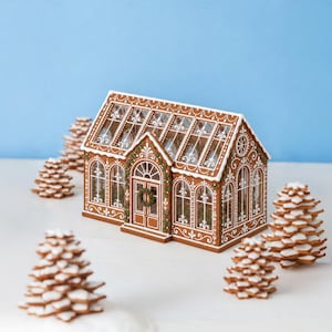 May include: A gingerbread greenhouse structure featuring brown walls with white icing details, a glass roof, and a small wreath on the front door. The structure is surrounded by several small, stacked cookie trees with white icing on the edges.