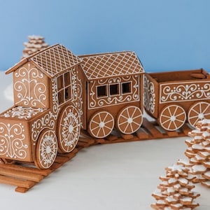 May include: A gingerbread train consisting of an engine, a passenger car, and a cargo car, all decorated with intricate white icing scrollwork and patterns. The train sits on gingerbread tracks, accompanied by two small stacked gingerbread trees.