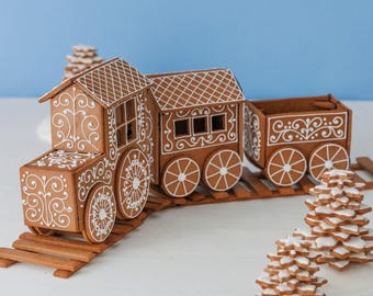 May include: A gingerbread train consisting of an engine, a passenger car, and a cargo car, all decorated with intricate white icing scrollwork and patterns. The train sits on gingerbread tracks, accompanied by two small stacked gingerbread trees.