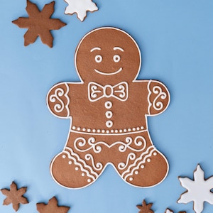 May include: A large gingerbread man cookie with white icing details, including a bow tie and buttons, surrounded by smaller gingerbread and snowflake cookies on a blue background. Festive holiday treats.
