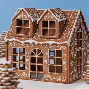 May include: A brown gingerbread house with white icing details, featuring two dormer windows on the roof, a central arched doorway, and multiple square window cutouts. The structure is decorated with white piped scrollwork and roof trim.
